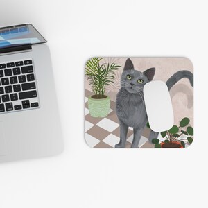 May include: A grey cat with green eyes sits on a chequered floor with two potted plants. The cat is looking to the left of the image. The background is a light pink colour.
