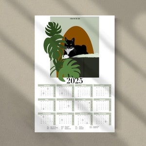 May include: A 2025 calendar featuring a black and white cat lounging on a brown and green surface. The cat is surrounded by green tropical leaves. The calendar is divided into 12 months, with each month displayed in a separate box.