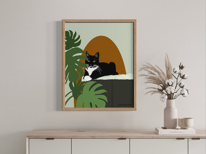 May include: A framed illustration of a black and white cat with green eyes, lounging on a ledge with a green monstera plant in the background. The illustration has a muted green, brown, and orange color palette. The text "FROM HERE" is at the bottom of the illustration.