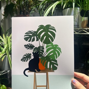 以下が含まれることがあります： 黒い猫が、オレンジ色の鉢植えのモンステラ植物の前にある木製の腰掛けに座っています。植物は大きく、緑の葉を持っています。背景は薄い青色です。画像は右下に「FROM RURU」と署名されています。