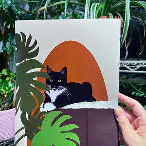 May include: A black and white cat with a white patch on its chest is lying on a brown surface with green leaves in the background. The cat is looking to the left of the image. The background is a light beige color with an orange arch.