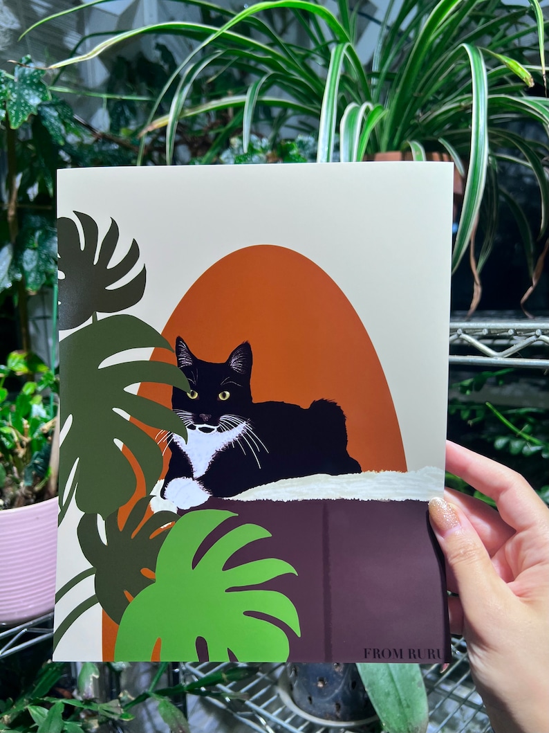 May include: A black and white cat with a white patch on its chest is lying on a brown surface with green leaves in the background. The cat is looking to the left of the image. The background is a light beige color with an orange arch.
