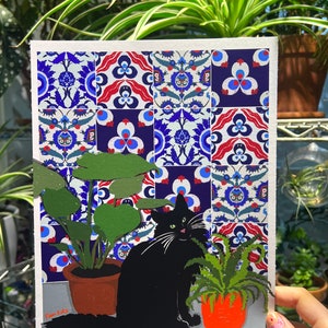 May include: A black cat sits in front of a tiled wall with blue, red and white geometric patterns. The cat is looking to the right. Two potted plants are on either side of the cat. The plant on the left has large, green leaves. The plant on the right has smaller, green leaves. The image is signed "Pam Katz".