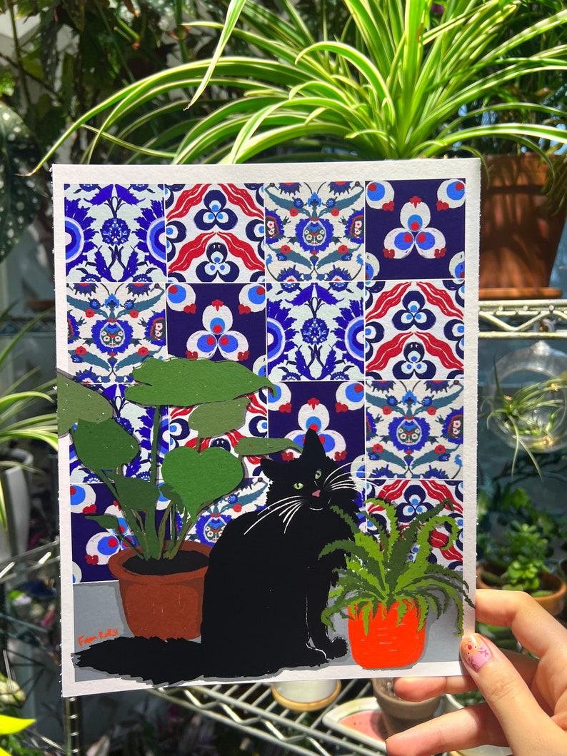 May include: A black cat sits in front of a tiled wall with blue, red and white geometric patterns. The cat is looking to the right. Two potted plants are on either side of the cat. The plant on the left has large, green leaves. The plant on the right has smaller, green leaves. The image is signed "Pam Katz".