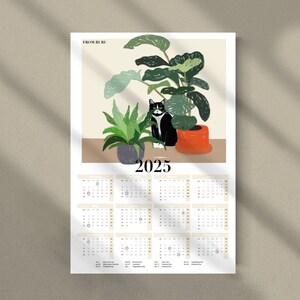 May include: A 2025 calendar featuring a black and white cat sitting in a pot of green plants. The calendar has a light beige background with black text. The top of the calendar has the text "From the Hill" in black.