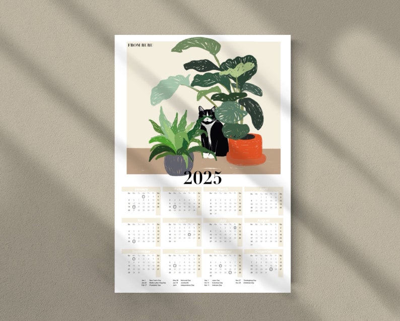 May include: A 2025 calendar featuring a black and white cat sitting in a pot of green plants. The calendar has a light beige background with black text. The top of the calendar has the text "From the Hill" in black.