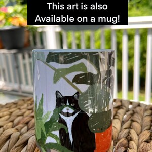 May include: White ceramic mug with a black and white cat illustration. The cat is peeking out from behind a green plant. The text "This art is also available on a mug!" is printed on the mug.
