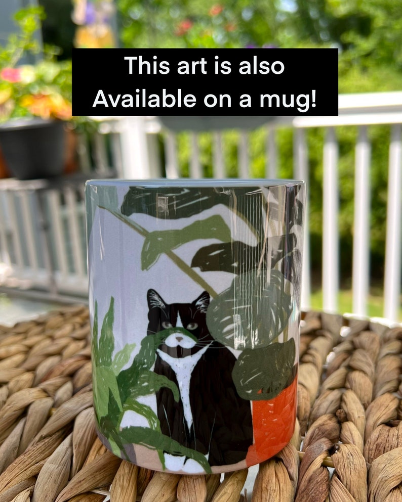 May include: White ceramic mug with a black and white cat illustration. The cat is peeking out from behind a green plant. The text "This art is also available on a mug!" is printed on the mug.
