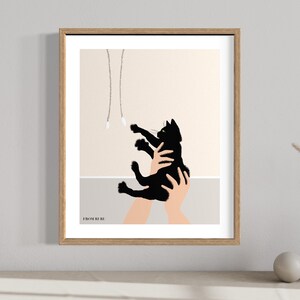May include: A framed art print featuring a black cat reaching for dangling toys. The cat is held in two hands, with a neutral background and a wooden frame. The print has a minimalist style.