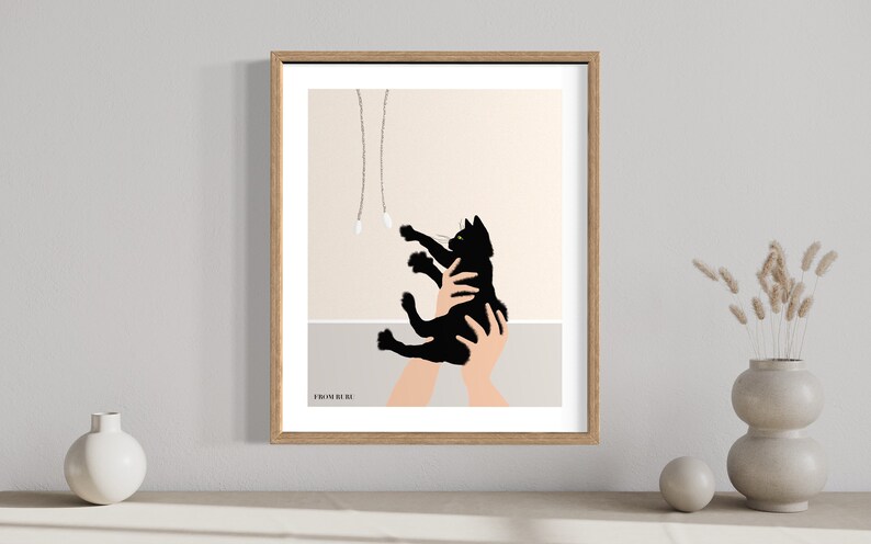 May include: A framed art print featuring a black cat reaching for dangling toys. The cat is held in two hands, with a neutral background and a wooden frame. The print has a minimalist style.