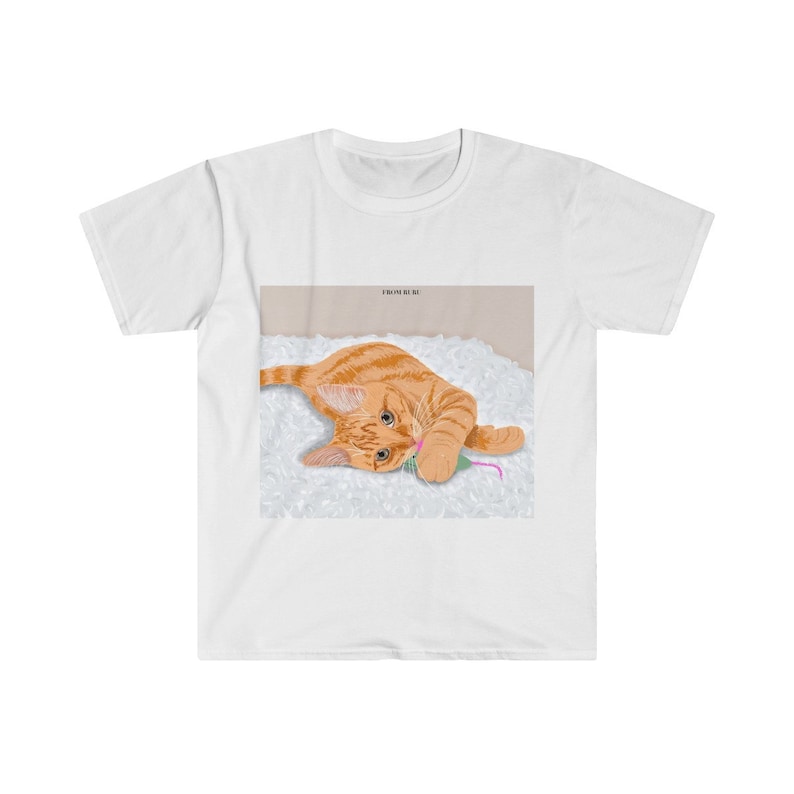 May include: A white t-shirt with a graphic of an orange tabby cat playing with a pink and green toy mouse on a white fluffy surface.