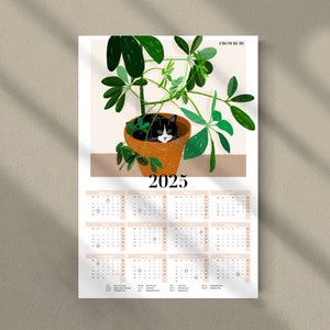 Pu&ograve; includere: Un calendario 2025 con un gatto bianco e nero rannicchiato in una pianta in vaso. La pianta ha grandi foglie verdi e il vaso &egrave; marrone. Il calendario &egrave; stampato su carta bianca con testo nero.