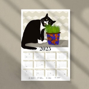 Peut inclure: Un chat noir et blanc aux yeux verts mange de l'herbe dans un pot bleu et orange. Le calendrier 2025 est situ&eacute; sous le chat. Le calendrier a un fond gris avec du texte noir.