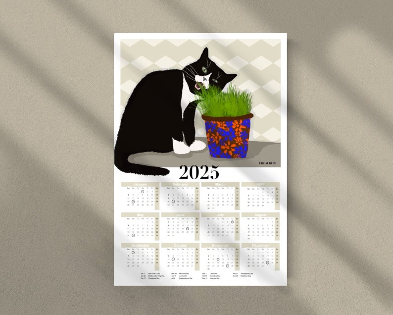 Peut inclure: Un chat noir et blanc aux yeux verts mange de l'herbe dans un pot bleu et orange. Le calendrier 2025 est situ&eacute; sous le chat. Le calendrier a un fond gris avec du texte noir.