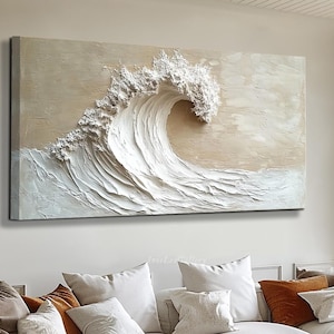 Puede incluir: Una pintura de lienzo texturizado blanco y beige de una gran ola rompiendo. La pintura está colgada en una pared sobre un sofá seccional blanco con almohadas.