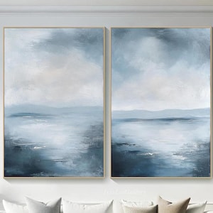 May include: Two abstract landscape paintings in blue and white tones, depicting a seascape with a horizon line and clouds. The paintings are framed in gold and hung on a white wall above a white sofa.
