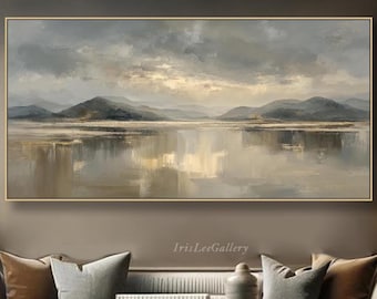Pode incluir: Pintura de paisagem emoldurada, retratando um lago com montanhas e reflexos, em tons de cinza, bege e dourado. A obra de arte está pendurada acima de um sofá em uma sala de estar. A moldura é dourada. A pintura tem formato retangular.