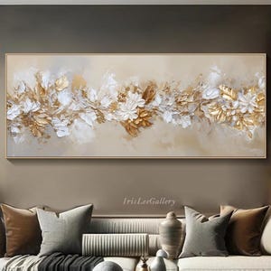 May include: A rectangular abstract painting in a gold frame. The artwork showcases a floral design in gold, white, and beige hues. The painting hangs above a white sofa with cushions, creating a luxurious living room setting.