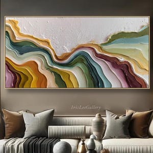 May include: A large, rectangular abstract painting with a gold frame. The artwork features layered, textured bands of color, including yellow, green, blue, and purple, creating a landscape effect. The painting hangs above a sofa in a living room.
