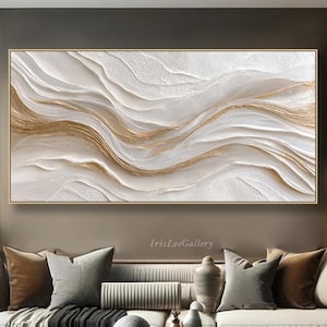 May include: A large, rectangular abstract painting with a gold frame. The artwork features a textured, layered design in white and gold, resembling flowing waves. The painting hangs above a sofa with decorative pillows, creating a modern living room aesthetic.