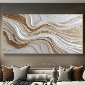Op de afbeelding: Een groot, rechthoekig abstract kunstwerk met een gouden lijst. Het kunstwerk heeft een textuurontwerp met vloeiende witte en gouden elementen. Het kunstwerk wordt boven een bank in een moderne woonkamer gepresenteerd.