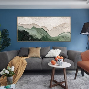 May include: A modern abstract landscape painting with a green and white color scheme. The painting is framed in a thin gold frame and hangs on a blue wall above a gray sofa.
