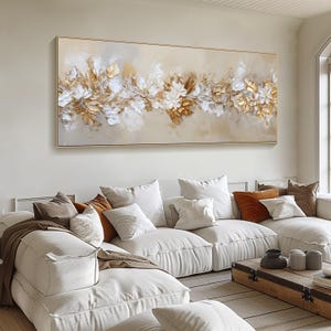 May include: A large rectangular canvas art piece featuring a floral design in gold, white, and beige tones. The artwork has a textured, three-dimensional appearance, with gold and white leaves and flowers. The artwork is framed in a thin gold frame.