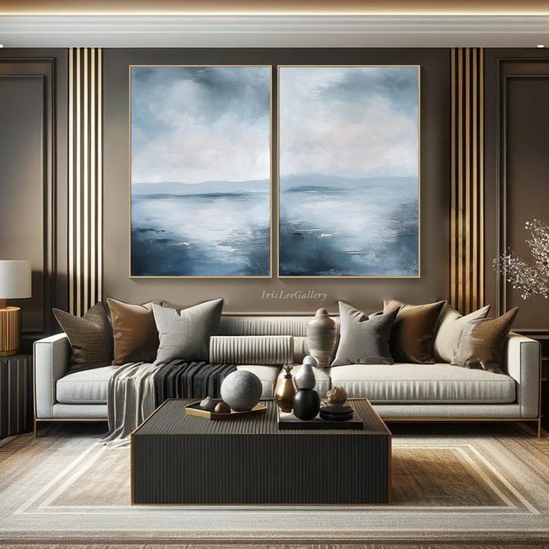 May include: Two abstract landscape paintings in shades of blue and white, hanging on a wall above a white sofa with brown throw pillows. The paintings are framed in gold.