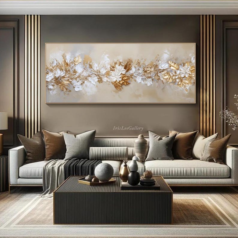 May include: A large, rectangular abstract painting with a gold frame. The artwork features a central floral design with gold, white, and beige tones. The painting is displayed above a white sofa with decorative pillows, creating a luxurious living room setting.