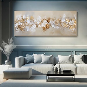 May include: A rectangular framed painting with a beige background. The artwork features a floral design with white and gold textured elements. The painting is displayed above a gray sofa in a modern living room setting.