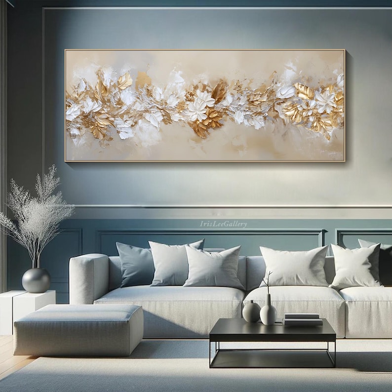May include: A rectangular framed painting with a beige background. The artwork features a floral design with white and gold textured elements. The painting is displayed above a gray sofa in a modern living room setting.