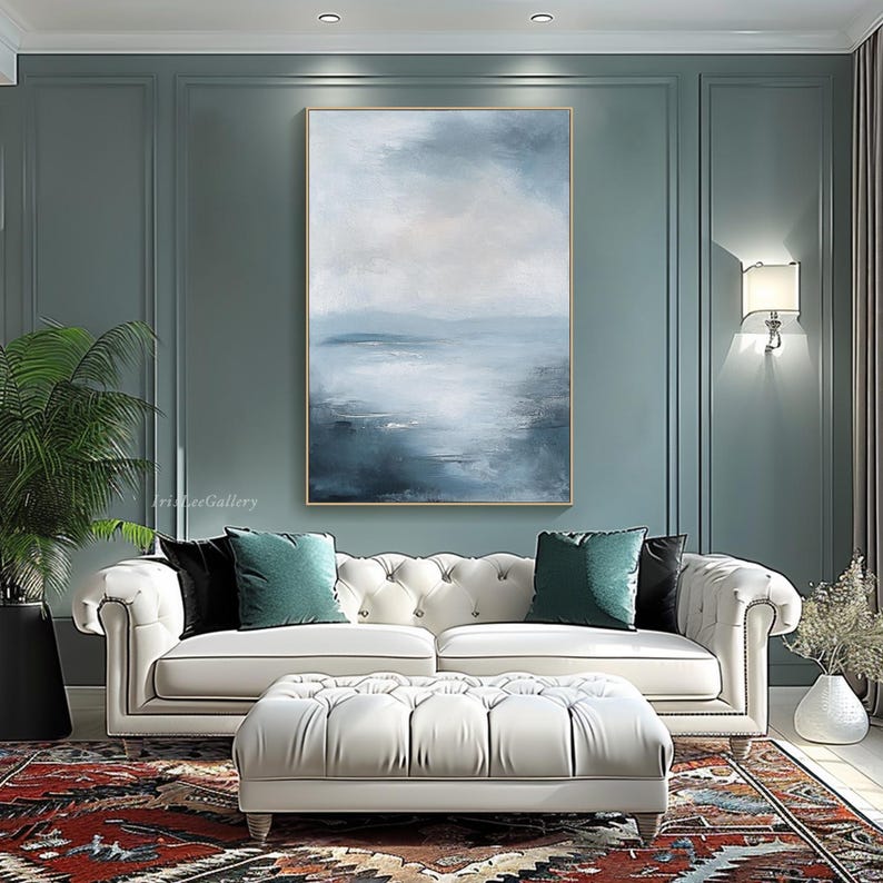 May include: A white sofa with tufted upholstery and two teal throw pillows sits in front of a large abstract painting of a blue and white seascape. The painting is framed in gold and hangs on a teal wall.