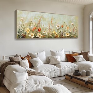 May include: A rectangular floral painting in a gold frame. The artwork features a textured, impressionistic style with yellow, orange, and white flowers against a pale green background. The painting is displayed above a white sectional sofa with various throw pillows.