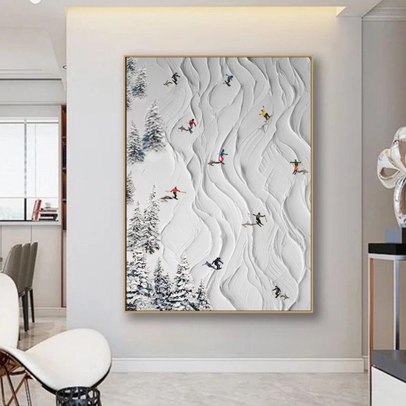 May include: A white and gray abstract painting with a textured surface. The painting depicts a snowy mountain scene with skiers and snowboarders in various colors. The painting is framed in a thin, gold frame.