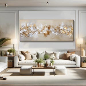May include: A large rectangular canvas art piece featuring a floral design with gold, white, and beige tones. The artwork is framed in a gold border and hangs above a white sofa in a living room setting.