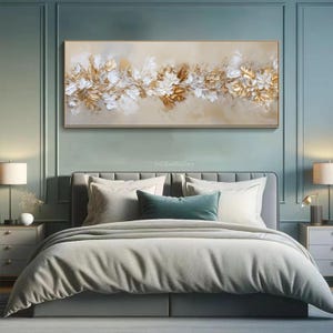 May include: A rectangular, framed artwork featuring a floral design in gold, white, and cream tones. The painting is displayed above a bed with a gray headboard, white pillows, and a gray comforter. The room has a teal-colored wall.