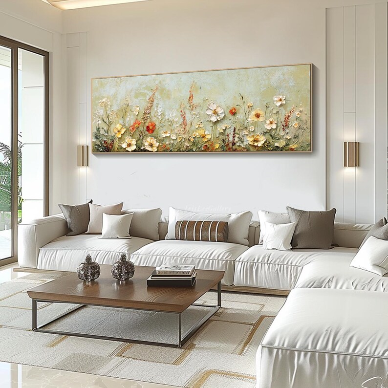 May include: A large rectangular floral painting with a gold frame hangs above a white sofa. The artwork features textured flowers in shades of yellow, orange, and white against a light green background. The room also includes a wooden coffee table and decorative pillows.