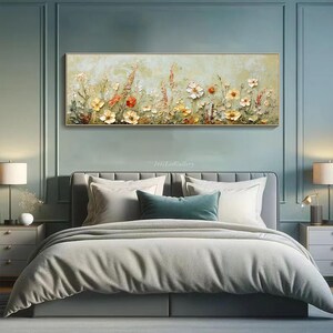 May include: A rectangular framed floral painting with a gold frame. The artwork features a textured, impressionistic style with white, yellow, and orange flowers. The painting hangs above a bed with gray bedding and white pillows.