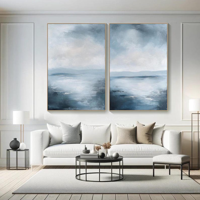 May include: Two abstract landscape paintings in blue and white tones, depicting a seascape with a horizon line and clouds. The paintings are framed in gold and hung on a white wall above a white sofa.