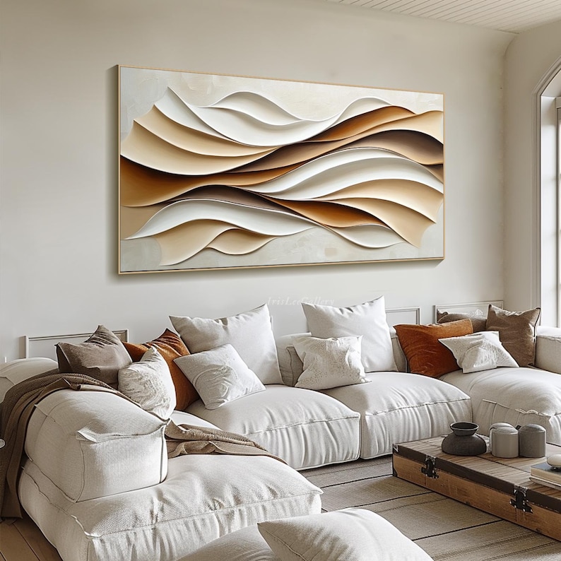 May include: Abstract wall art featuring a layered design of white, brown, and beige waves. The artwork is framed in a thin gold frame and hangs on a white wall above a white sectional sofa.