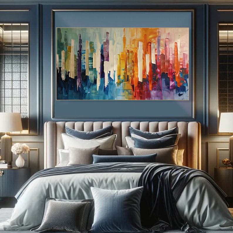 Puede incluir: Pintura abstracta de un horizonte urbano con rayas verticales de colores en tonos de azul, morado, amarillo, naranja y rojo. La pintura est&aacute; enmarcada y colgada en una pared sobre una cama con ropa de cama azul y blanca.
