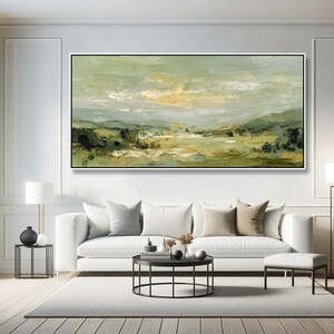 May include: An abstract landscape painting in a black frame. The artwork features a palette of greens, yellows, and whites, depicting a serene countryside scene. The painting hangs above a white sofa with neutral-toned pillows.