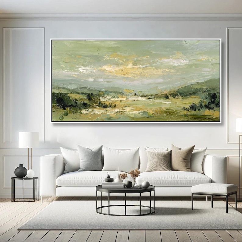 May include: An abstract landscape painting in a black frame. The artwork features a palette of greens, yellows, and whites, depicting a serene countryside scene. The painting hangs above a white sofa with neutral-toned pillows.