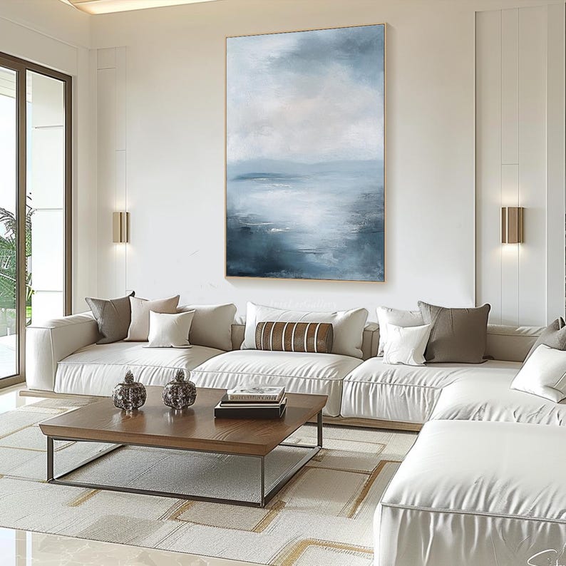 May include: A white sectional sofa with a brown and white striped throw pillow in front of a large abstract painting of a blue and white seascape. The painting is hanging on a white wall in a modern living room.