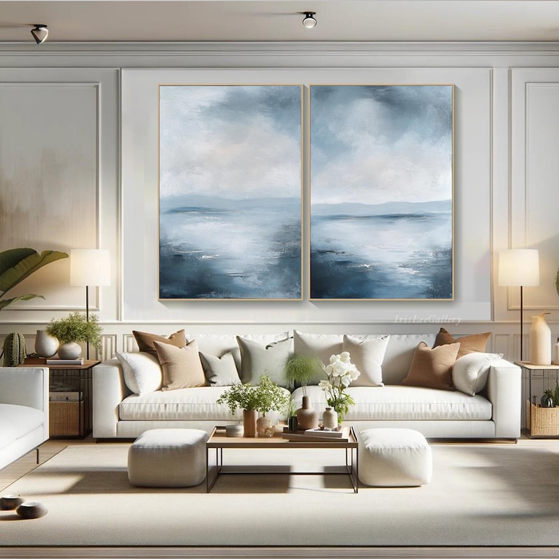 May include: A set of two abstract landscape paintings in shades of blue and white, depicting a seascape with a horizon line and a cloudy sky. The paintings are framed in gold and hung on a white wall above a white sofa.