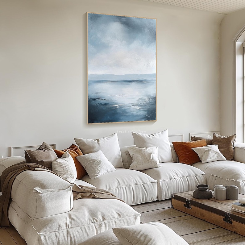 May include: A large abstract painting of a seascape with a blue and white color scheme. The painting depicts a calm ocean with a distant horizon and a cloudy sky. The painting is hanging on a wall in a living room setting.