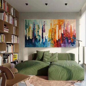 Puede incluir: Pintura abstracta de ciudad con pinceladas verticales de colores en tonos azules, amarillos, naranjas, rojos y morados. La pintura est&aacute; colgada en una pared sobre un sof&aacute; verde con cojines.