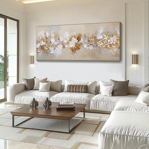 May include: A large rectangular abstract painting with gold, white, and beige floral elements. The artwork is framed in a gold-colored border and hangs above a white sofa. The room has a modern, minimalist design.