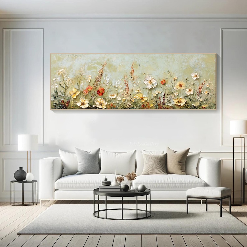 May include: A rectangular floral painting with a gold frame. The artwork features a textured, three-dimensional depiction of various flowers in shades of yellow, orange, and white against a light green background. The painting hangs above a white sofa in a modern living room.