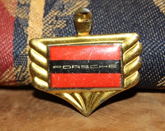 May include: A gold-colored Porsche emblem with a red and black rectangular center. The word "PORSCHE" is printed in white across the black stripe. The emblem has a decorative, ridged border and a loop at the top.